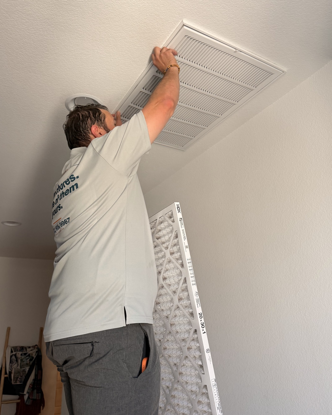 A technician in a Saltbox polo lifting the grille off a hallway ceiling return, a new filter propped against the wall below